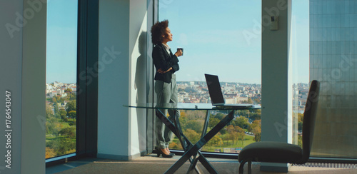 Obraz na plátně Young businesswoman with curly hair standing by a large office window, holding a