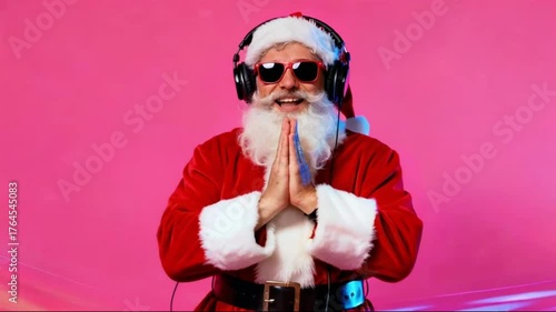 A full shot of Santa Claus wearing sunglasses and headphones, dancing with his fingers pointing upward against a bright pink background.