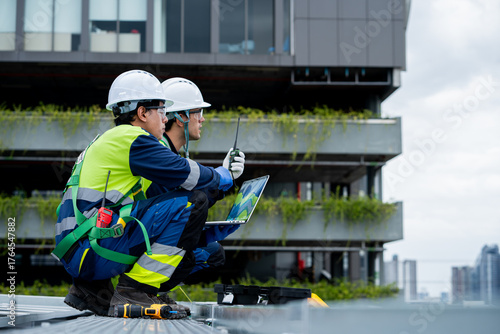 Two men in safety gear are looking at a laptop on a rooftop
