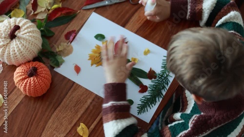 Sweet child, boy, applying leaves using glue while doing arts and crafts at home, autumntime