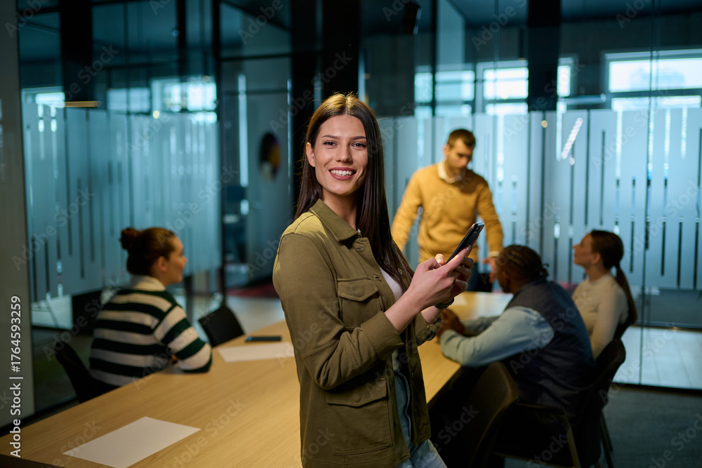 Naklejka premium Confident business woman smiling while using smartphone during a team meeting in a modern office environment. Concept of communication, leadership, and professional confidence.