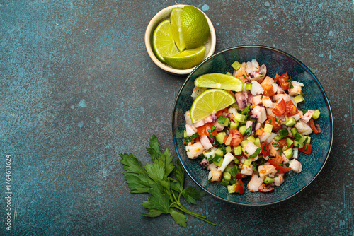Fresh and vibrant peruvian seafood ceviche with shrimps, octopus, tomatoes, cucumber and parsley, served with lime in a bowl on a stone background from above, dish of Peru. Copy space