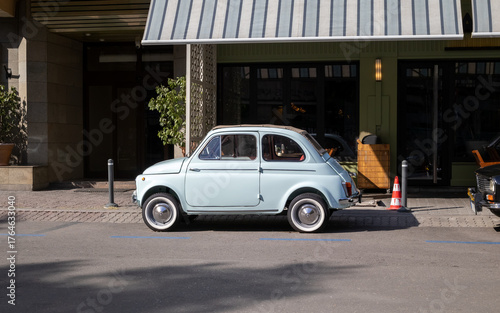 Fototapeta Naklejka Na Ścianę i Meble -  side view of a small, light blue vintage Fiat 500 car parked on a sunny city street in front of a modern cafe or shop