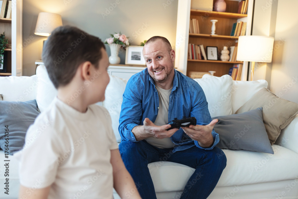 Obraz premium Father and son play video games with joystick and have fun at home. Father get angry after he lose match and happy son game winner