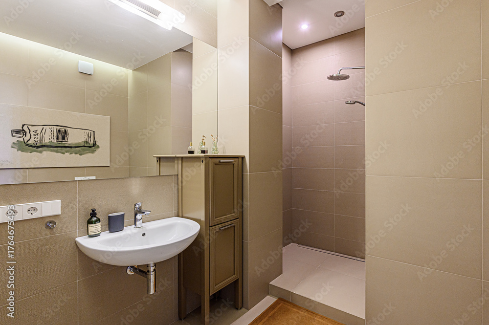 Obraz premium Modern bathroom with beige tile walls, a white sink, and a walk-in shower. A mirror reflects artwork above the sink