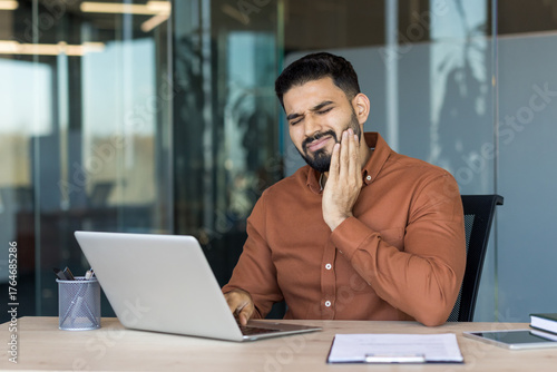 Young man experiencing dental pain while working on his laptop in a modern office, stopping work due to the discomfort and needing to visit a dentist for relief
