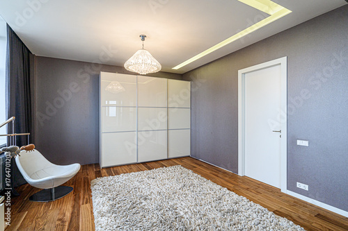 A modern bedroom showcases a white closet, stylish chair, soft rug, and wood floor. Gray walls add contrast