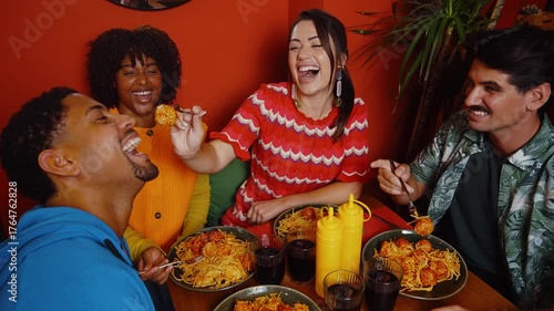 Happy group of diverse friends having fun while eating spaghetti and meatballs together in a colorful restaurant, with a laughing woman playfully feeding her male friend a meatball