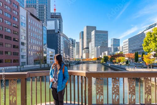 Asian woman solo traveler tourist holding a camera, standing on a bridge, and enjoying the modern cityscape view of Osaka, Japan on a sunny autumn day.