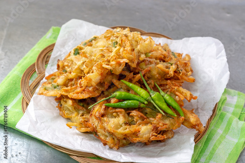 A stack of crispy Indonesian bakwan sayur (vegetable fritters) served on white paper with a group of green chili peppers on a woven bamboo plate.