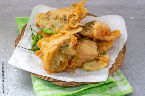 A small stack of Indonesian tahu isi (deep-fried stuffed tofu) with a crispy batter coating and green chili peppers on a woven bamboo plate.