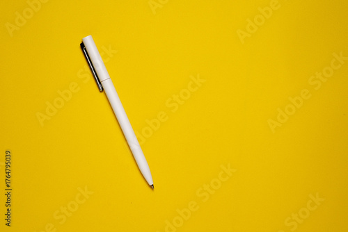 A top-down view of a single white ballpoint pen with a silver clip, placed diagonally on a bright yellow textured background.