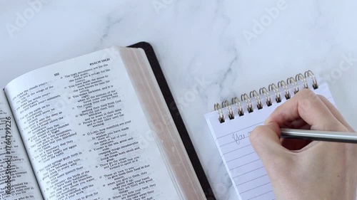 Human hand is writing inspirational christian text in a notebook while studying from holy bible book. Top view. Biblical concept.