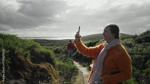 Woman in an orange sweater takes a picture with her smartphone while walking along a scenic coastal path.