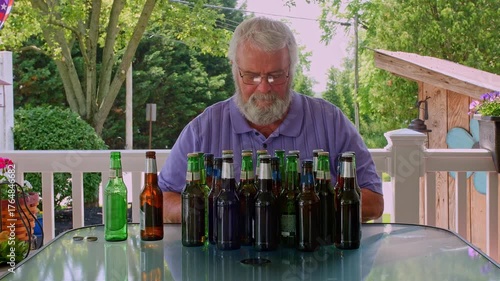 An elderly man sits at a table on his patio, smiling while surrounded by an assortment of empty beer bottles. The sunlight creates a warm atmosphere as he reflects on his day.