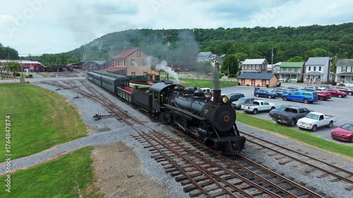 A vintage steam train pulls out of the station, chugging down the tracks in a small town surrounded by green hills. The scene captures the charm of rail travel and the beauty of the area.