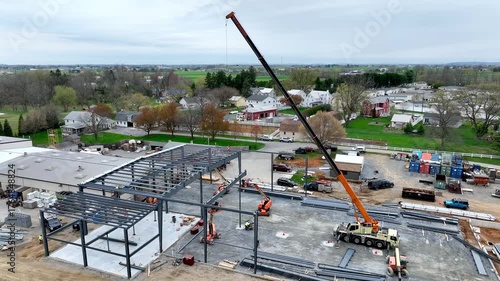 Large construction site shows a crane lifting steel beams into place. Workers coordinate below as structures take shape in the countryside. Green fields surround the area.