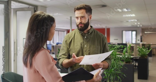 Consulting male wearing green shirt showing printed papers to female in office, with black folder