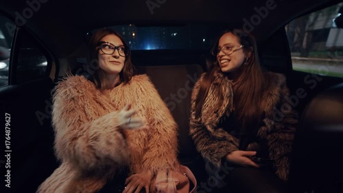 Two stylish girls in luxurious fur coats sit in the back seat of a taxi, laughing and chatting as the bright city lights flash through the windows at night.
