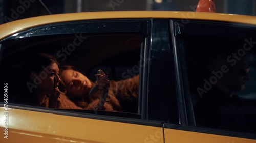 Glamorous women enjoy a taxi ride through the city at night, taking photos and doing makeup in the back seat while bright city lights flicker through the car window.