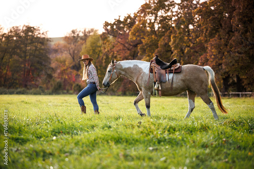 Cowgirl in western clothing leading horse across pasture during autumn sunset. Woman walking with horse, symbolizing slow living and country lifestyle
