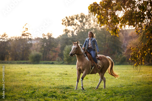 Fotografie Woman riding horse through autumn pasture at sunset