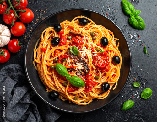 Classic Spaghetti alla Puttanesca, an Italian pasta dish with tomatoes, black olives, capers, and basil, served in a black bowl on a dark background.