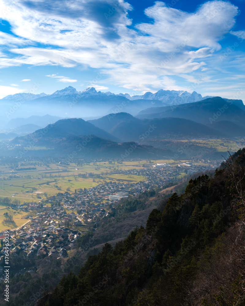 Fototapeta premium Misty mountains overlook a village and valley under blue skies.