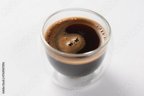 A transparent double-layer glass coffee cup filled with rich dark brown liquid, featuring a dense layer of fine foam on top with tiny bubbles clinging to the inner wall