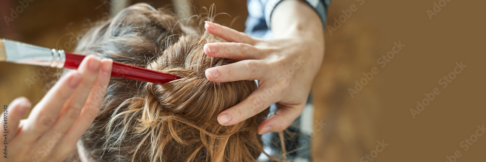 Fototapeta premium Female applying hair dye with brush on brown hair close-up in hair salon setting.