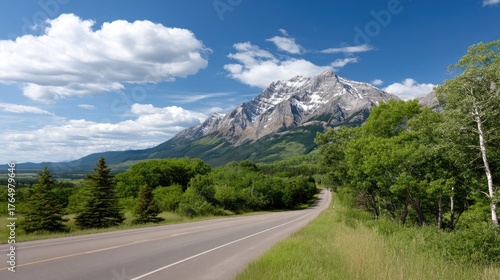 Cars travel on a winding road beside a tranquil lake surrounded by snowy mountains under a vibrant blue sky with clouds