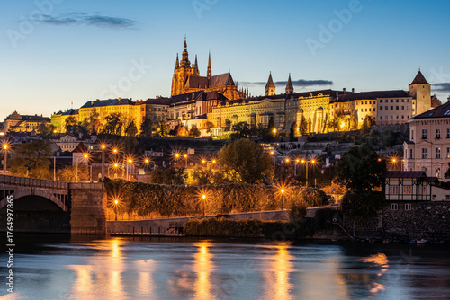 Prague Castle illuminated at blue hour with reflections on the Vltava River and city lights glowing in the evening sky.