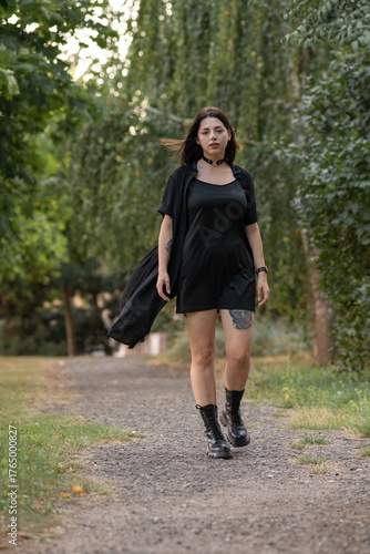 Female model in black clothing poses confidently on a forest trail, holding outfit straps, with vibrant greenery creating a serene and stylish atmosphere