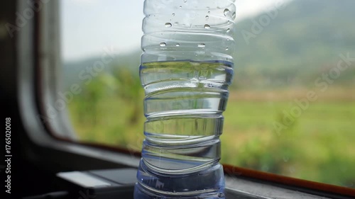 Close-up of a plastic water bottle on a train window with a scenic green landscape in the background.