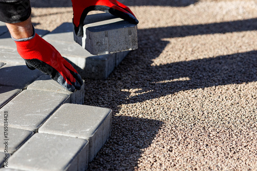 Worker laying concrete paving blocks for sidewalk construction, Construction worker with red gloves placing gray concrete paving stones on gravel surface for new pavement or walkway installation.