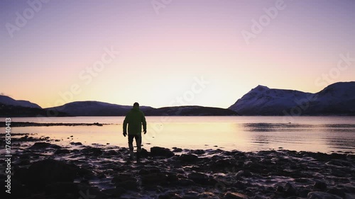 Man in winter clothing walks thoughtfully across ice crusts on the shore of the Atlantic Ocean during an icy winter in Northern Norway near Tromso