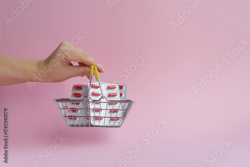 A hand holds a shopping basket filled with blister packs of pills against a bright pink background with space for text, symbolizing the growing cost of pharmaceuticals