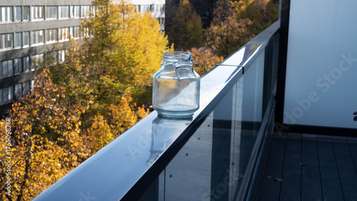 A broken glass jar on a balcony railing, with yellow autumn trees below.
