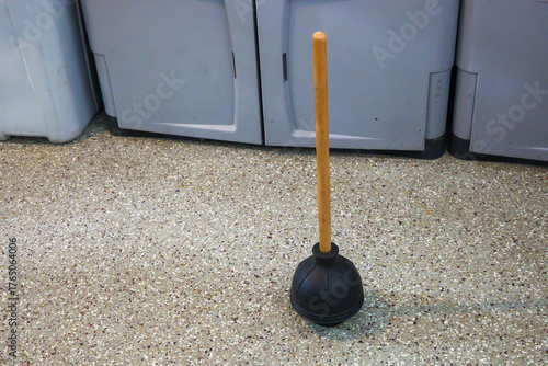 A black rubber toilet plunger with a wooden handle standing upright or vertical on the epoxy covered garage floor.