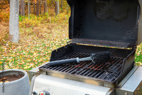 An open gas grill with a metal cleaning brush on the grates, outside in autumn with leaves on the lawn.