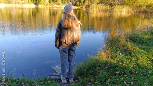 A girl in a warm jacket and a knitted hat stands on the shore of a lake.