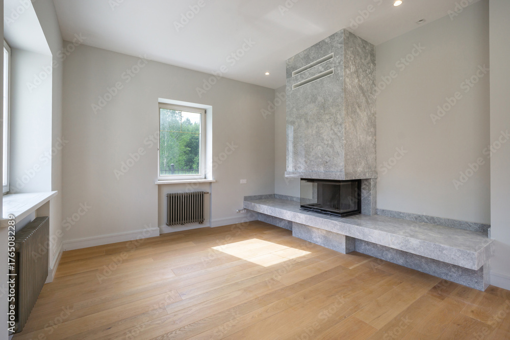 Fototapeta premium A bright living room features a sleek, stone fireplace and large windows that allow natural light to fill the space. The wooden floor adds warmth to the minimalist decor.