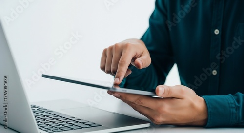 A close-up shot of a man in a dark green shirt using a tablet while sitting in front of a laptop. The focus is on his finger tapping the screen against a clean, white background.