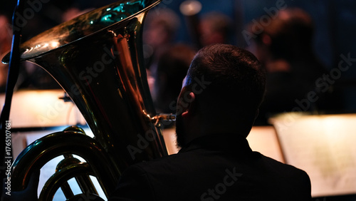 Rear view of brass player in silhouette holding french horn during concert with blurred musicians and warm stage lights in background 