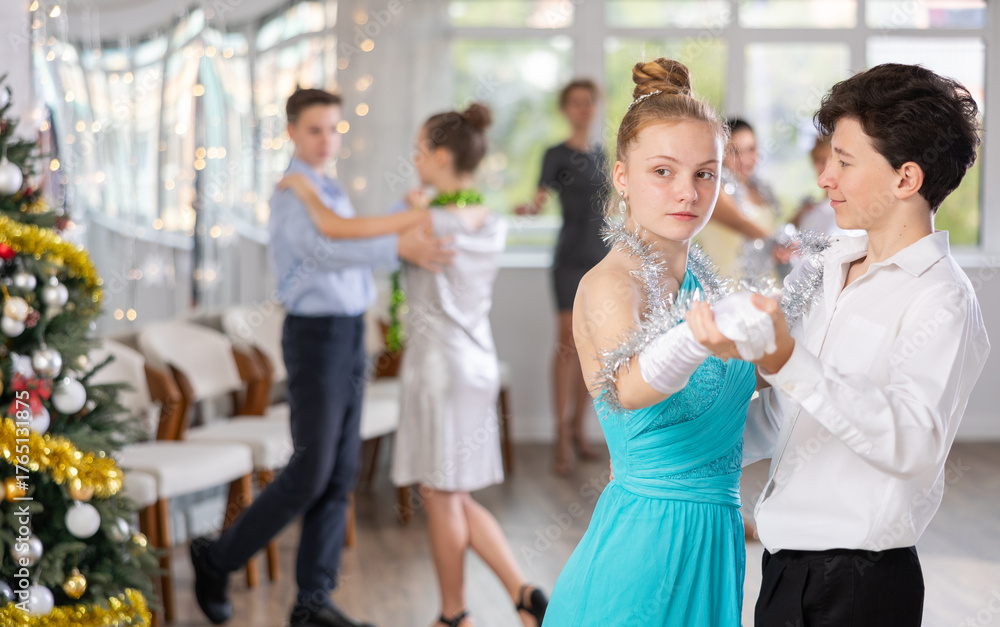 Fototapeta premium Positive teenage students in festive attire rehearsing ballroom dance number for upcoming Christmas party at college with interested female teacher in background..
