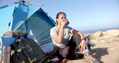 Sitting adult woman sipping from metal cup at coastal bluff campsite, with blue tent and backpack