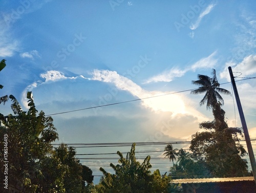 A view of the sky with a large, dramatic cloud formation, with the sun's rays shining through. The foreground features silhouettes of tropical trees, including a palm tree, and power lines.