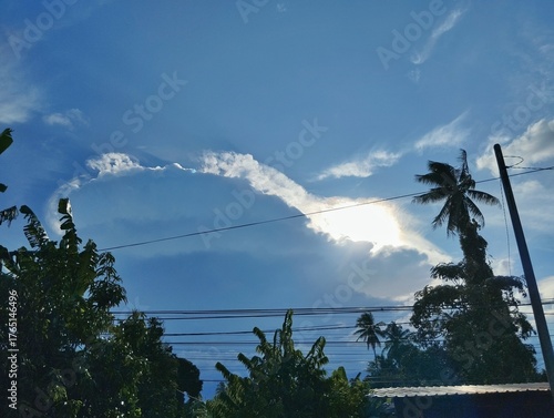 A view of the sky with a large, dramatic cloud formation, with the sun's rays shining through. The foreground features silhouettes of tropical trees, including a palm tree, and power lines