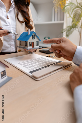 A real estate agent and client discussing property sale details at the office table, reviewing and signing home purchase contracts with miniature house models and money on the desk.
