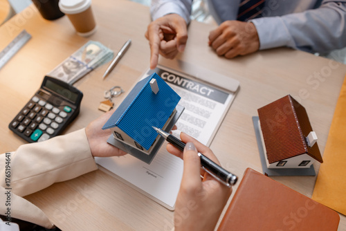 A real estate agent and client discussing property sale details at the office table, reviewing and signing home purchase contracts with miniature house models and money on the desk.
